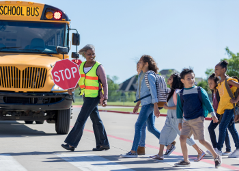 Student children crossing the street to enter a school bus guided by an adult holding a stop sign and wearing a safety vest